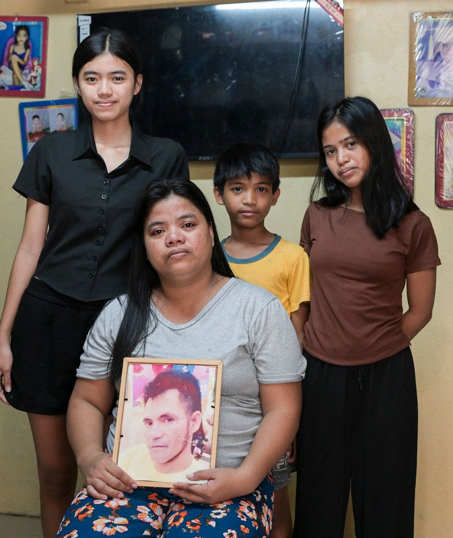 Drei Frauen und ein Mann aus der indigenen Bevölkerungsgruppe in Mindanau auf den Pilippinen sitzen in bunten, folkloristischen Kleidern auf einer Holzbank vor einem Gebäude aus Bambus. Ein kleines Kind steht neben einer der sitzenden Frauen und sieht ebenfalls in die Kamera.