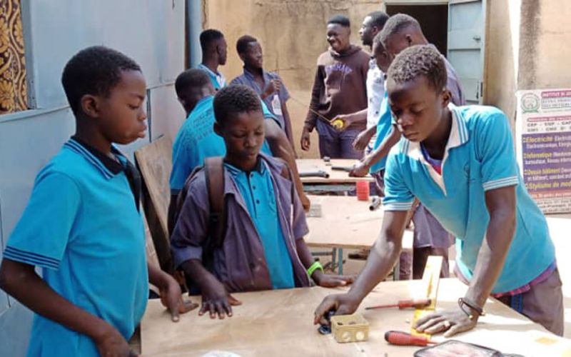 Jugendliche in Burkina Faso erlernen ein Handwerk: im Vordergrund stehen drei Jugendliche in hellblauen T-Shirts an einer Werkbank.