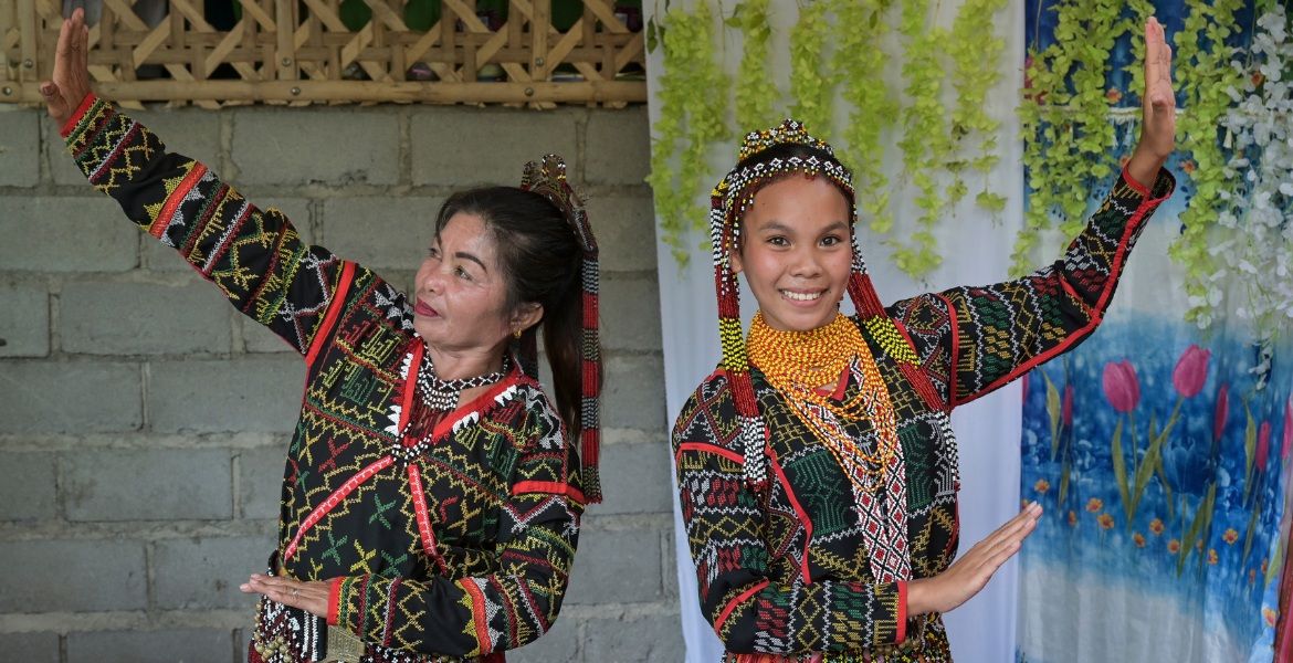 Eine jüngere und eine ältere Frau aus der Provinz Cotabato auf den Philippinen zeigen in ihrer traditionellen Kleidung einen Tanz, bei dem sie sich spiegelsymmetrisch zueinander bewegen.