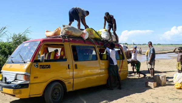 Das Dach eines gelben Kleinbusses wird in Madagaskar auf sandigem Boden von mehreren Männern mit Säcken beladen.