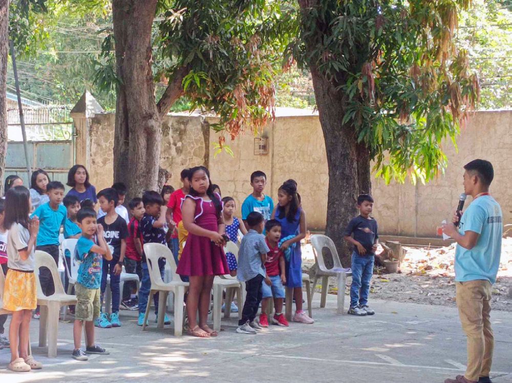 Ein angehender Katechist steht auf den Philippinen in einem Innenhof mit einem Mikrofon vor einer Gruppe von Kindern.
