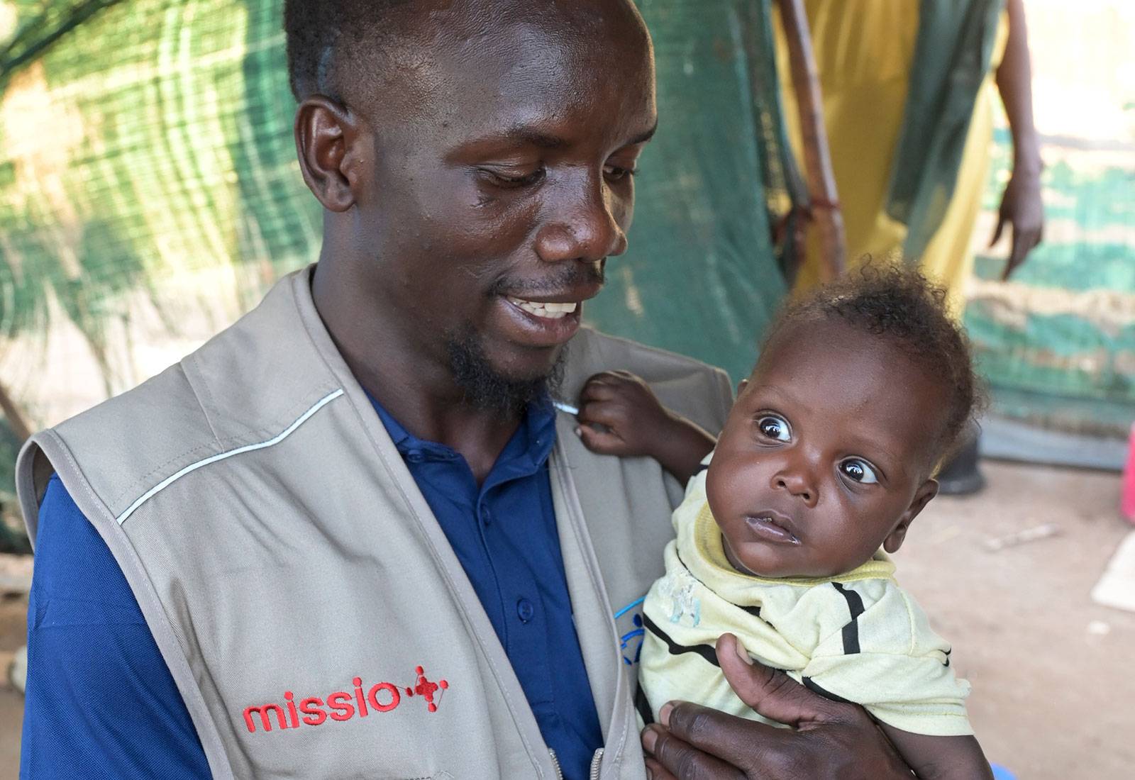 Ein Helfer vom JRS mit missio-Logo auf seiner Weste hält ein Baby in den Armen, das verschreckt guckt.
