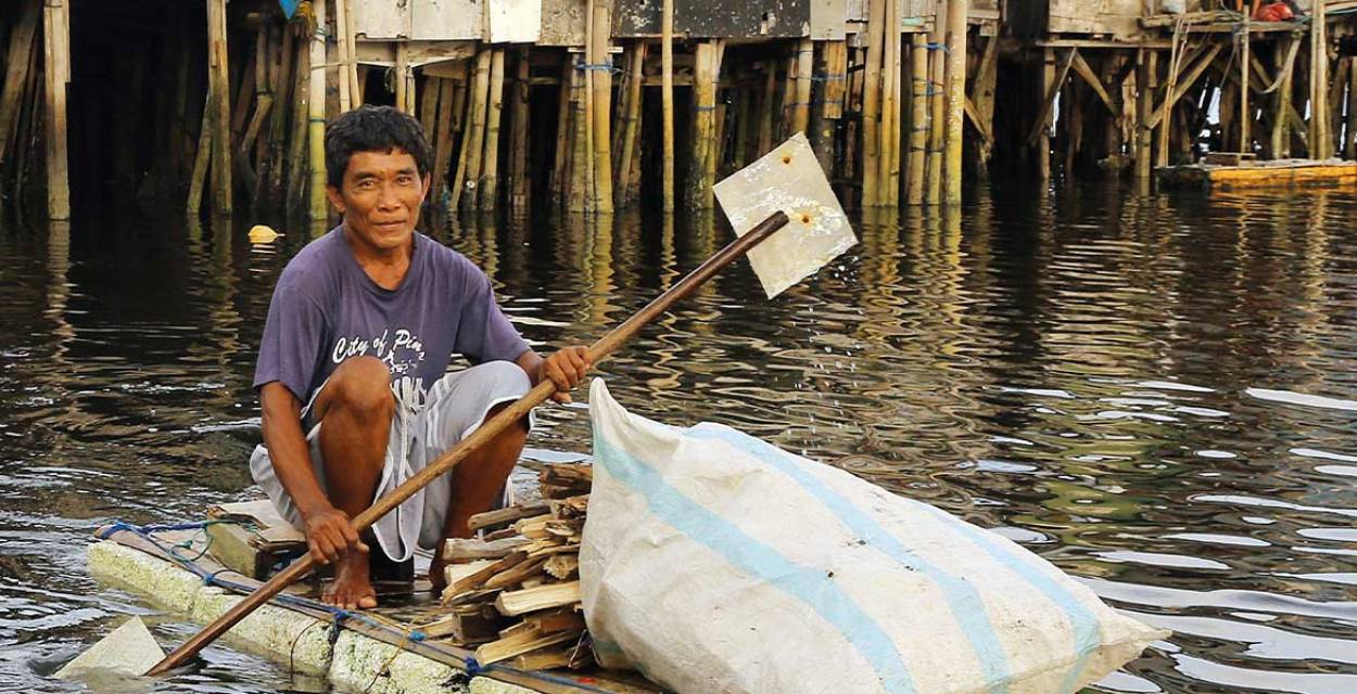Ausschnitt aus dem missio-Plakat zum Weltmissionsmonat 2025: Ein Mann paddelt auf einem Floß vor Stelzenhäusern in Manila.