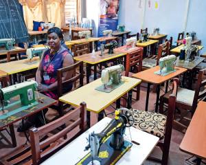Eine Frau sitzt allein im Schulungsraum, vorne eine Tafel mit Schnittmustern, auf den Tischen stehen Nähmaschinen. Davor je ein leerer Stuhl.