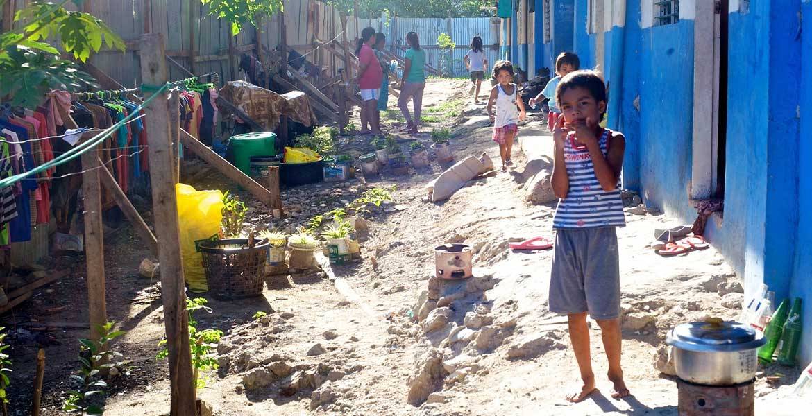 In einem Armenviertel von Cebu auf den Philippinen steht ein Kind im Vordergrund. Vor ihm auf dem Boden ein Topf auf einer offenen Feuerstelle, Kleider hängen auf Kleiderstangen auf der Seite des Weges.