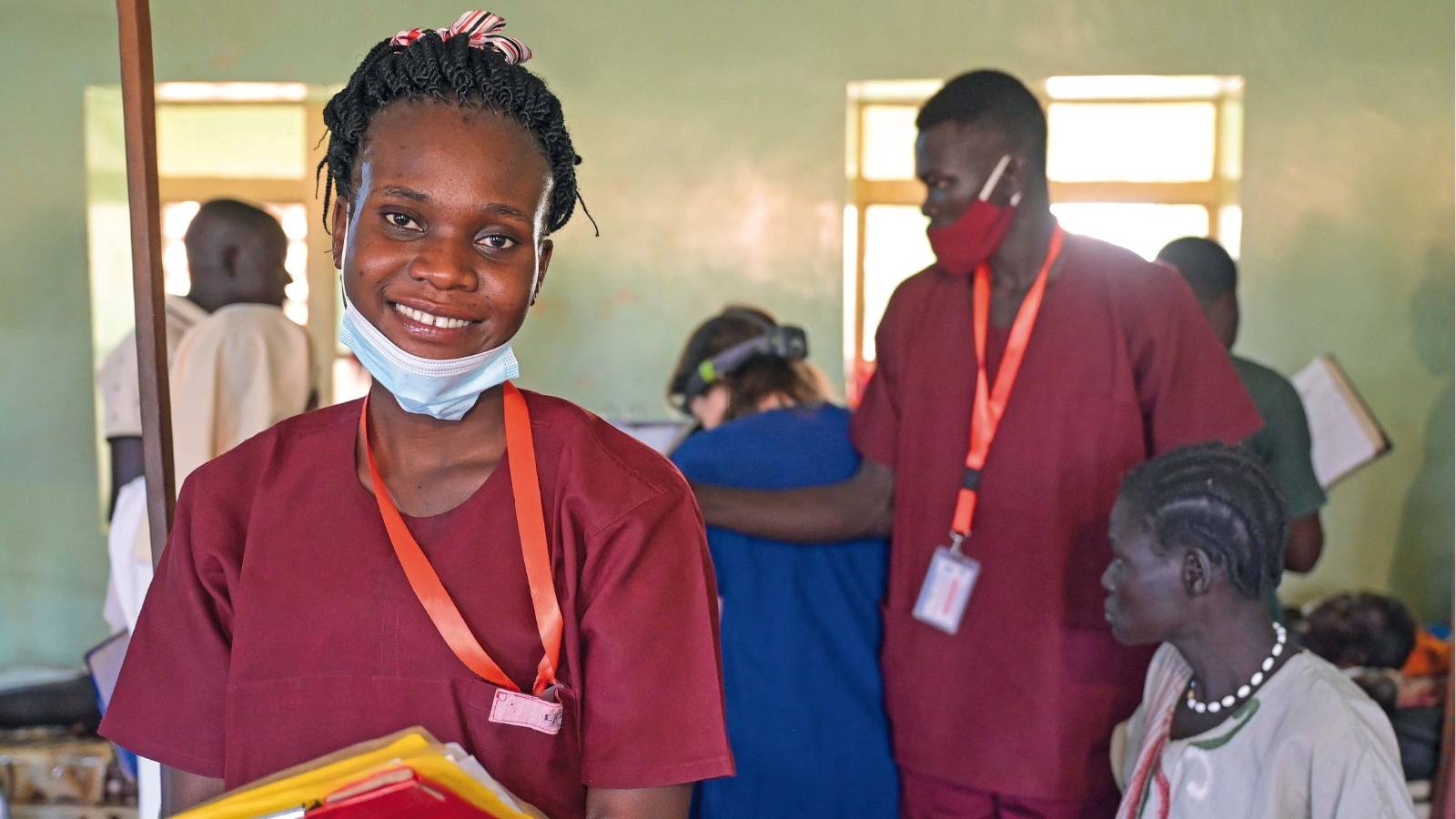 Links im Bild: die angehende Hebamme Edia in ihrer bordeauxroten Arbeitskleidung. Sie hat die OP-Maske heruntergezogen und lächelt. Im Arm hält sie Patientenakten. Im Hintergrund rechts von Edia: ein anderer Angestellter des Krankenhauses sowie weitere Personen, vielleicht Patienten.