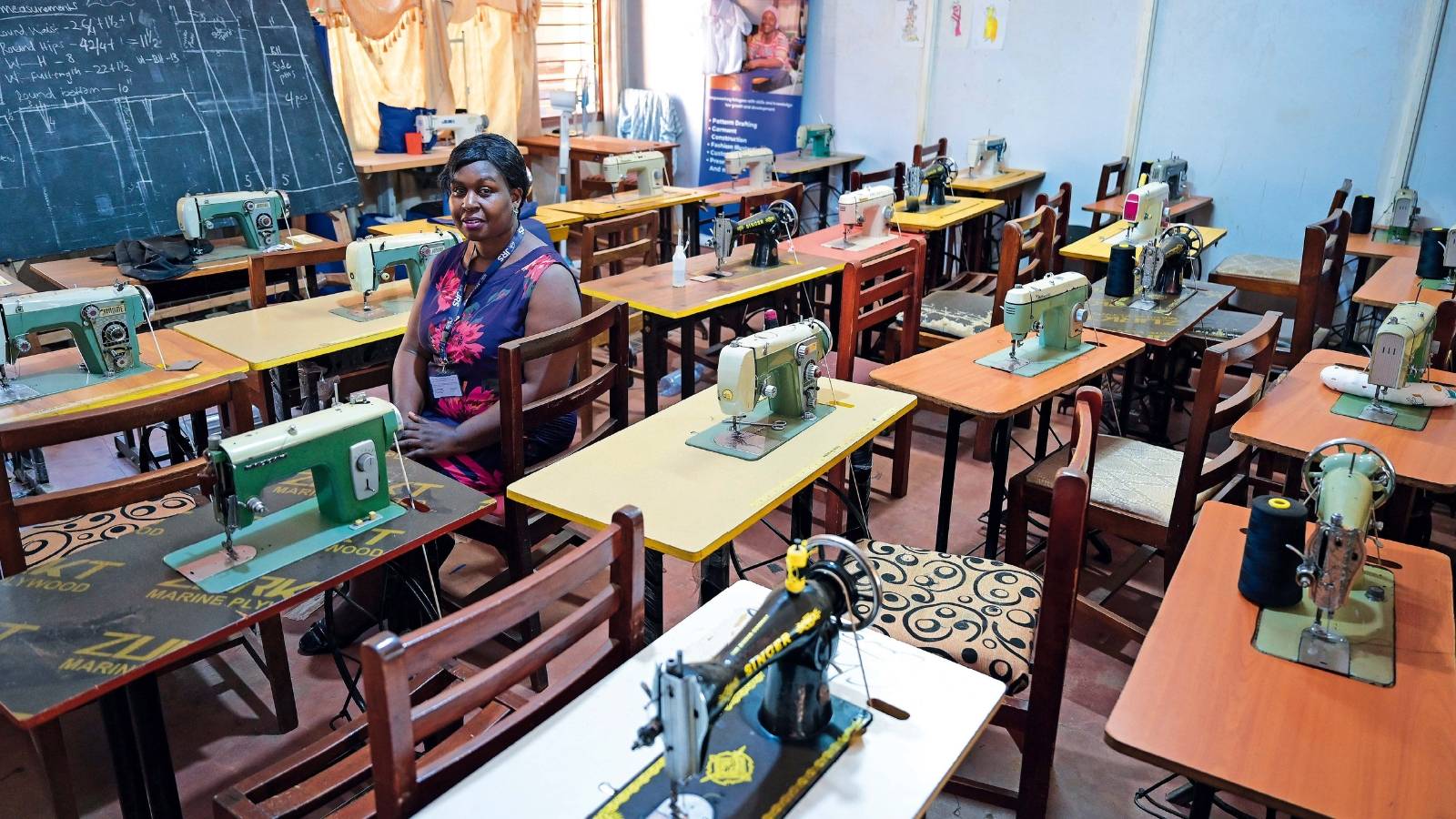 Eine Frau sitzt allein im Schulungsraum, vorne eine Tafel mit Schnittmustern, auf den Tischen stehen Nähmaschinen. Davor je ein leerer Stuhl.