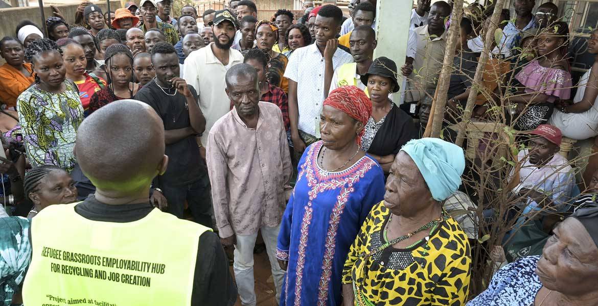 Ein Ordner steht (mit dem Rücken zur Kamera) in einer neongelben Weste in Kampala/Uganda vor einer Gruppe von geflüchteten Menschen aus dem Kongo.