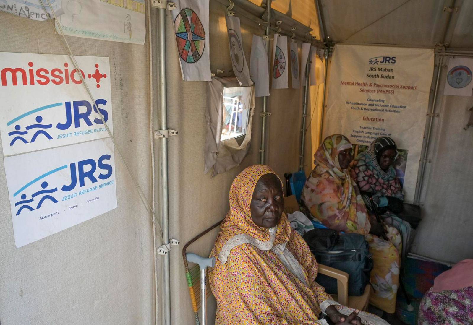 drei Frauen mittleren Alters mit bunten Tüchern bekleidet sitzen neben einer Tür mit der Aufschrift missio - Jesuit Refugee Service und warten.