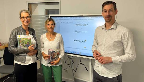 Zwei Mitarbeiterinnen von der Benefizaktion Sternstunden stehen links vor einem großen Monitor, rechts davon steht missio-Fundraiser Christian Homey. Alle drei blicken in die Kamera.