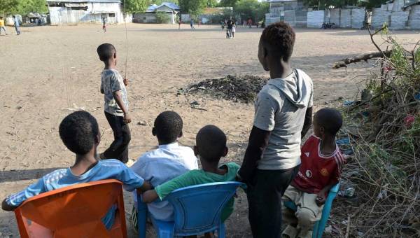 Vier Kinder sitzen im Halbschatten auf Plastikstühlen. Zwei weitere stehen. Sie wenden der Kamera den Rücken zu und blicken auf einen weitläufigen, sandigen Platz.