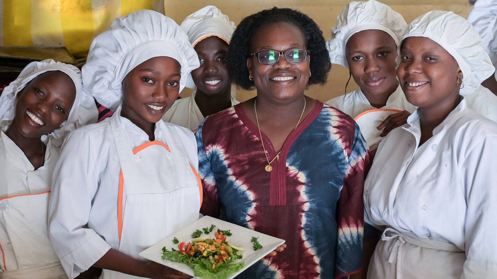 Junge Frauen stehen lächelnd in weißer Küchenkleidung um Louise Ndione, Leiterin der gemeinnützigen Ausbildungseinrichtung Claire Amitié in Thies, Senegal. Marie in der Mitte präsentiert einen Vorspeisensalat.