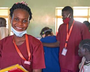 Links im Bild: die angehende Hebamme Edia in ihrer bordeauxroten Arbeitskleidung. Sie hat die OP-Maske heruntergezogen und lächelt. Im Arm hält sie Patientenakten. Im Hintergrund rechts von Edia: ein anderer Angestellter des Krankenhauses sowie weitere Personen, vielleicht Patienten.