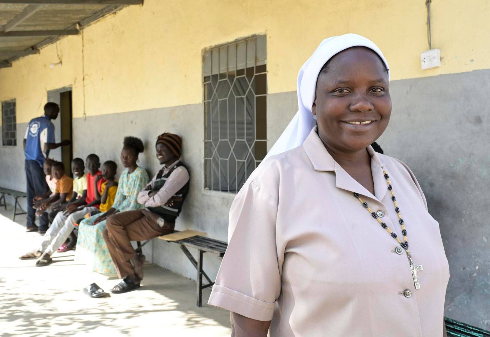 Schwester Amony Susan Atube läuft außen an einem Genbäude vorbei und lächelt in die Kamera. Hinter ihr sitzen auf einer Bank mehrere Kinder und zwei junge Erwachsene.