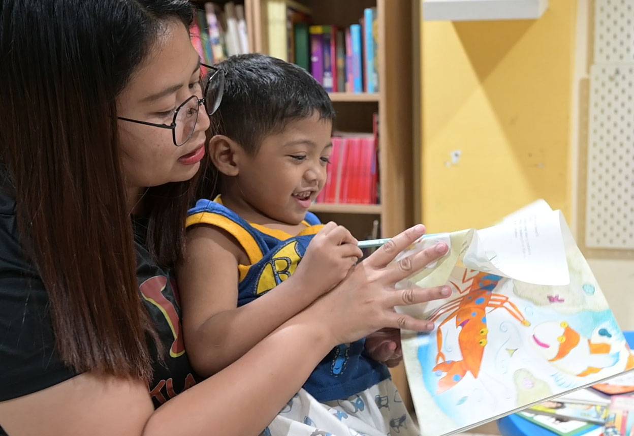 Eine Betreuerin hat einen kleinen Jungen auf dem Schoß, sie schauen ein Heft mit Bildern an
