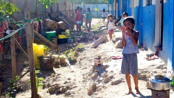 In einem Armenviertel von Cebu auf den Philippinen steht ein Kind im Vordergrund. Vor ihm auf dem Boden ein Topf auf einer offenen Feuerstelle, Kleider hängen auf Kleiderstangen auf der Seite des Weges.