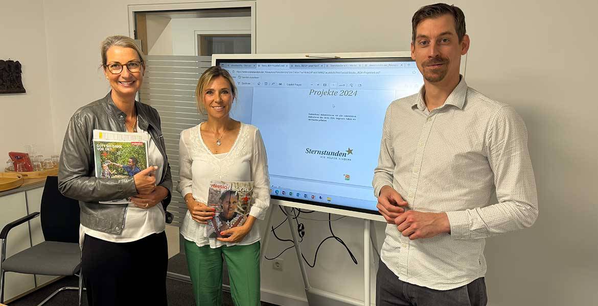 Zwei Mitarbeiterinnen von der Benefizaktion Sternstunden stehen links vor einem großen Monitor, rechts davon steht missio-Fundraiser Christian Homey. Alle drei blicken in die Kamera.